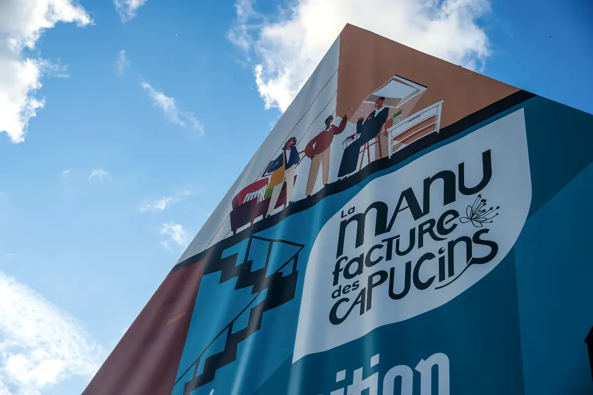Vue en contre-plongée du haut de la fresque de la Manufacture des Capucins, montrant trois personnages et le grand logo sur fond bleu.